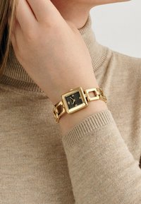 Gold rectangular watch on wrist, featuring a black face with white hour markers, linked gold metal band, worn over a beige textured sweater.