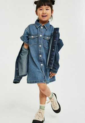 Jong meisje dat glimlacht, gekleed in een knoopjurk van denim en een bijpassend denim jasje vasthoudt, met lichte sneakers en patroon sokken.