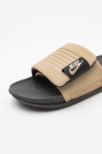 Beige és fekete Nike papucs, párnázott pánttal, rajta a Nike logóval, fehér háttéren.