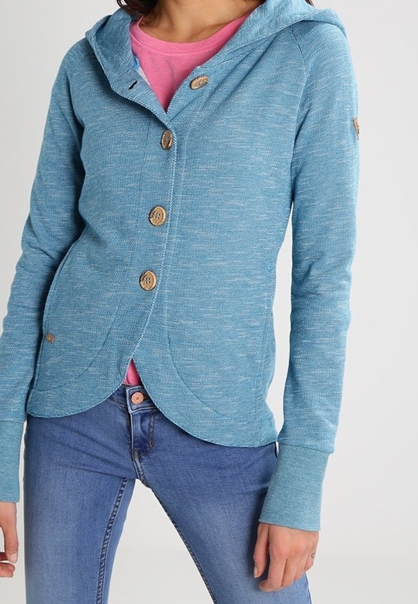 Femme portant un cardigan à capuche bleu boutonné par-dessus un t-shirt rose et un jean bleu sur un fond uni.