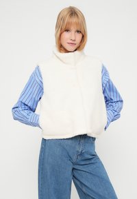 Bela flisasta brezrokavnik z visokim ovratnikom, oblikovan preko modre črtaste srajce, kombiniran z modrimi jeans hlačami. Mehka tekstura in skrajšana zasnova.