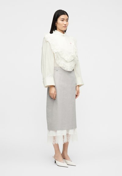 Γυναίκα που στέκεται φορώντας λευκό πουκάμισο με δαντελένια βολάν, ανοιχτόγριμη μίντι φούστα, εσώρουχο με δαντέλα και λευκές μυτερές γόβες.