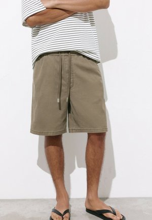 Homme portant un short kaki à cordon, une chemise blanche à rayures noires et des tongs noires, debout les bras croisés contre un mur blanc.