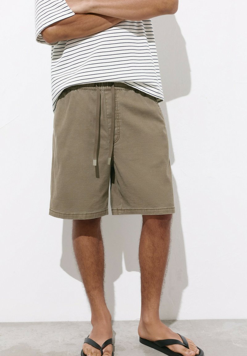 Homme portant un short kaki à cordon, une chemise blanche à rayures noires et des tongs noires, debout les bras croisés contre un mur blanc.