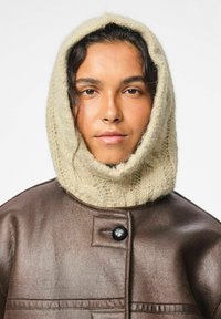 Kvinde iført en beige strikket hætte og en brun læderjakke med en stor sort knap, som ser direkte ind i kameraet.