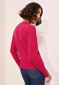 Pulover ajustat, cu mânecă lungă, din tricot roz strident, cu margine cordonată. Prezintă mansete texturate cu trei butoane decorative. Gât clasic de tip crew.