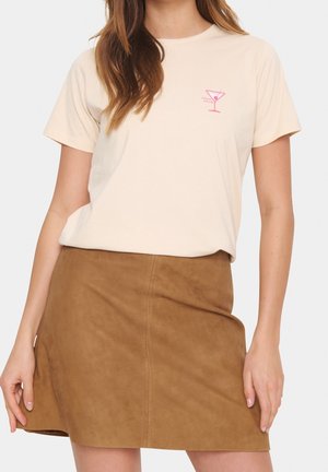 Femme portant un t-shirt beige avec un petit motif de verre à cocktail rose et une mini-jupe en daim marron.