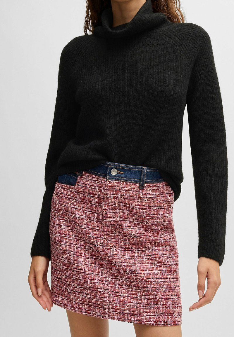 Kvinna klädd i en svart stickad polotröja och en texturerad röd minikjol med detaljer i denim vid midjan.