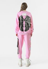Conjunto de sudadera y pantalones rosa. La sudadera presenta un diseño de alas de lentejuelas negras en la parte posterior; los pantalones están decorados con acentos bordados en blanco y dorado.