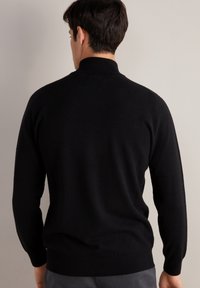 Svart tröja med hög krage, långa ärmar, mjuk textur och ribbad nederkant. Designen betonar en figursydd siluett och sömlös konstruktion.