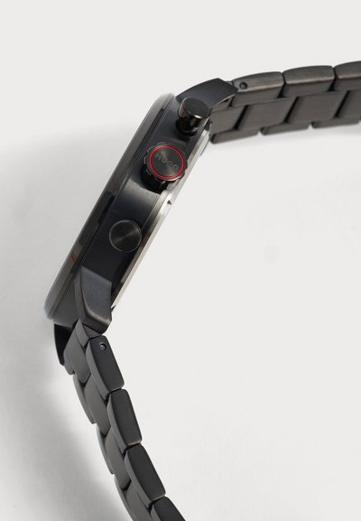 Vue latérale d'une montre-bracelet en métal noir montrant trois boutons ronds, un avec anneau rouge et gravure "HUGO", et bracelet à maillons.