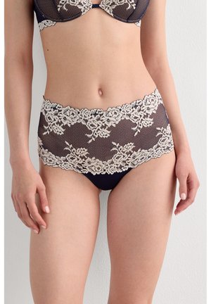 Frau trägt marineblaues Dessous-Set mit beigen floralen Spitzenverzierungen am BH und hoch tailliertem Slip, stehend vor weißer Wand.