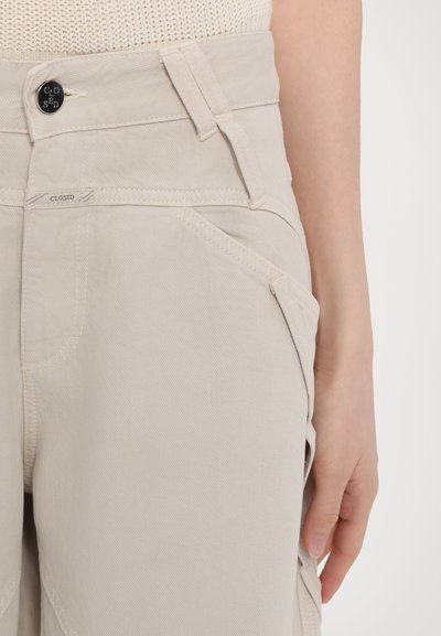 Beige denim pants with a high waist, featuring angled front pockets and a closed button. The fabric has a textured finish.