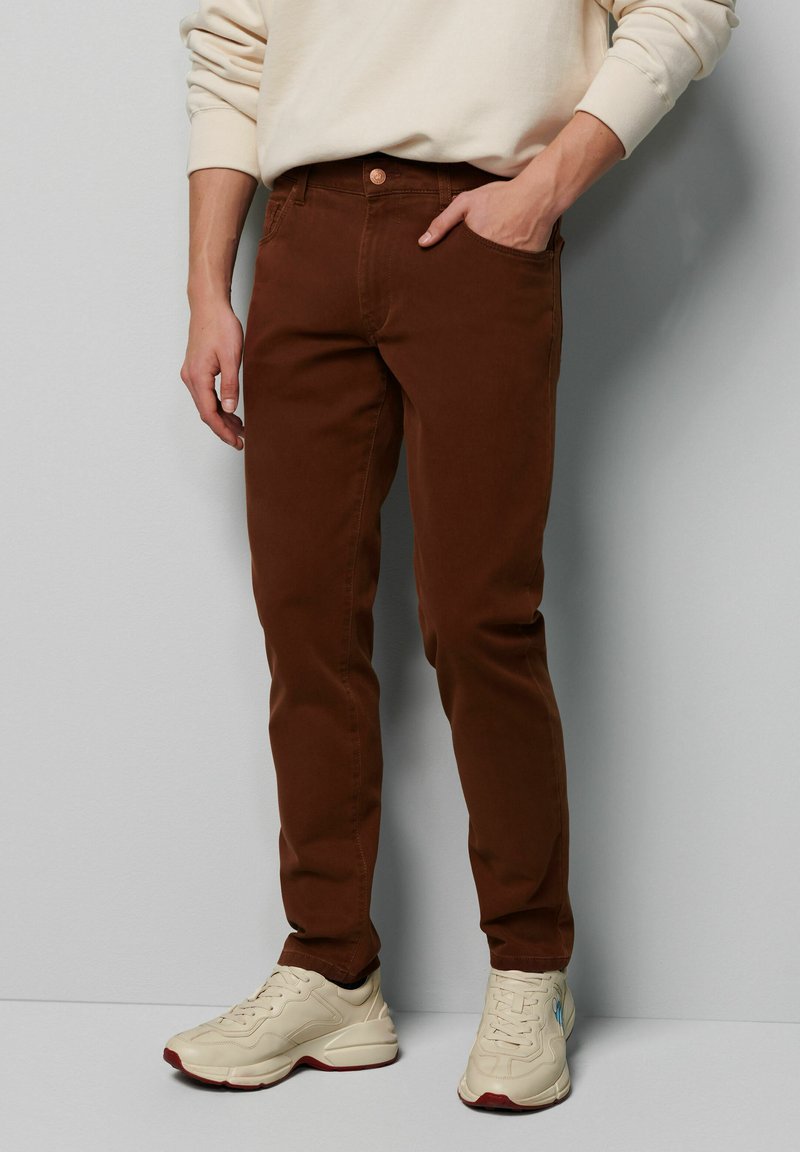 Braune Baumwoll-Skinny-Jeans mit einer Fronttasche, einem Metallknopf und gesteppten Nähten, kombiniert mit beigen Sneakers. Die Textur wirkt glatt.