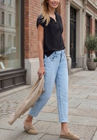 Frau geht auf einer Straße in der Stadt, trägt hellblaue Jeans, ein schwarzes kurzärmeliges Oberteil, beige Slipper und hält eine beige Jacke in einer Hand.