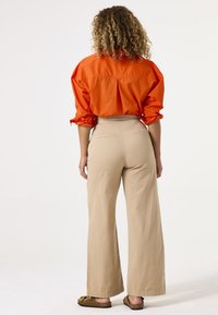 Oranje katoenen shirt met opgerolde mouwen, gecombineerd met lichtbeige wijde broekspijpen. Schoeisel zijn bruine sandalen met decoratieve hardware.