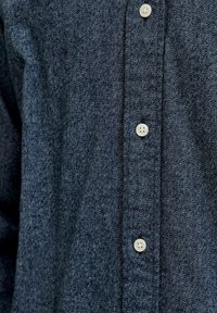 Tuvumā redzama tumši zila teksturēta auduma krekls ar trim redzamām baltām pogām, kas aizpogātas priekšā.