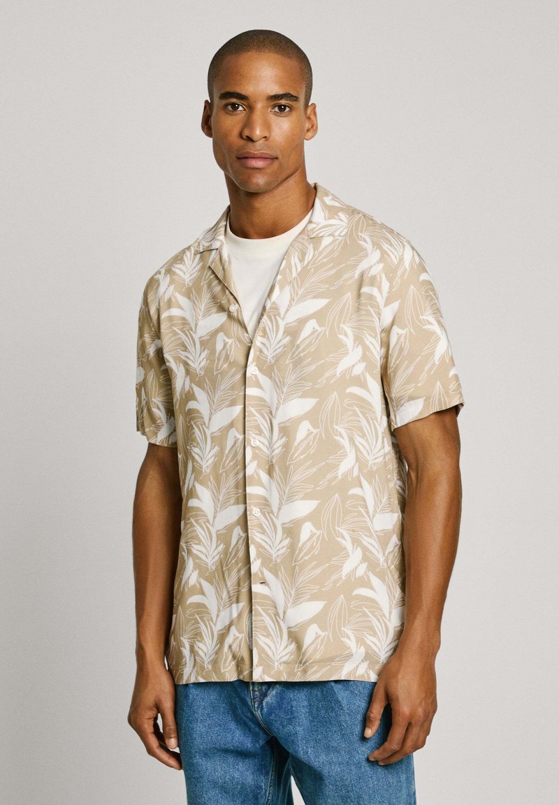 Camicia a maniche corte beige con motivo a foglie bianche, caratterizzata da un colletto a cappello e chiusura con bottoni, abbinata a jeans blu.