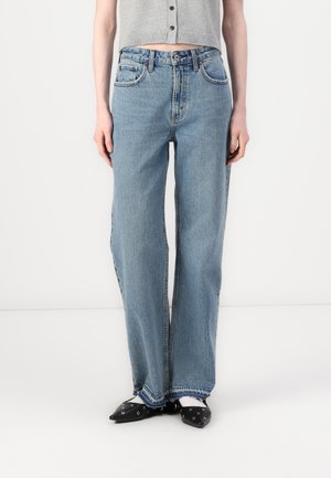 Personne portant un jean large bleu clair aux ourlets bruts, un gilet gris court boutonné et des chaussures noires pointues avec des œillets argentés.