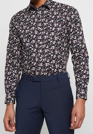 Man draagt een zwart overhemd met lange mouwen en kleine witte bloemenprint, gecombineerd met donkerblauwe nette broek.