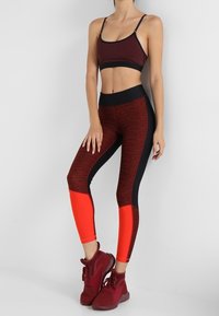 Zestaw sportowy składający się z biustonosza sportowego i legginsów w kolorze burgundowym z czarnymi akcentami. Legginsy mają jasnoczerwone dolne panele oraz teksturowany materiał. Dobrane do tego czerwone buty sportowe.