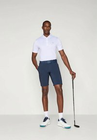 Hombre con camiseta polo blanca y pantalones cortos azul marino sostiene un palo de golf, vistiendo calcetines blancos y zapatos de golf blancos con suelas azules.