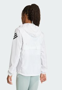 Biela bunda s kapucňou, so čiernym trojprúžkovým dizajnom na rukáve a sťahovacím lemom. Hladká, ľahká tkanina.