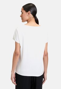 Biała bluzka z krótkim rękawem o luźnym kroju, okrągłym dekolcie, gładkim materiale i lekko zaokrąglonym dole, noszona z czarnymi, luźnymi spodniami.