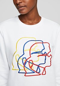 Sudadera blanca con contornos de rostros bordados en colores rojo, azul y amarillo, con un cuello redondo y una textura suave.
