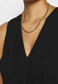 Zlatý řetízek s proplétanými články, se lesklým povrchem a hladkou strukturou, nošený přes černý top s výstřihem do V.