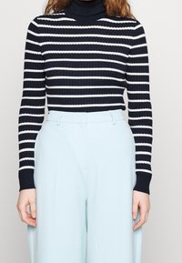 Granatowy i biały prążkowany sweter z wysokim kołnierzem, połączony z jasnoniebieskimi spodniami z wysokim stanem. Miękka tekstura i dopasowany krój.