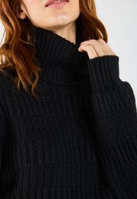 Maglione a collo alto nero a costine con un collo spesso e maniche lunghe. La texture sembra morbida e lavorata a maglia, evidenziando un materiale caldo.