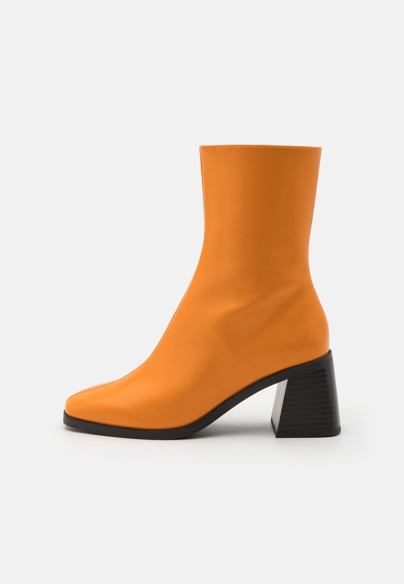 Botín naranja hecho de material suave con punta afilada y tacón geométrico negro. El diseño es elegante y ajustado.