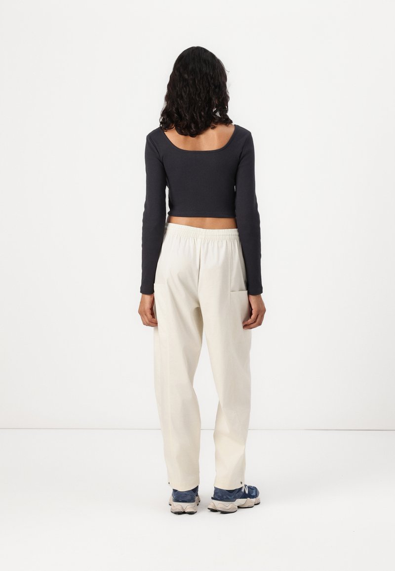 Haut court à manches longues de couleur sombre avec un décolleté rond, associé à un pantalon large crème et des chaussures de sport bleues, se tenant devant un fond uni.