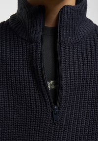 Jūras spektra adīts pulovers ar augstu apkakli un rāvējslēdzēju, ar strukturētu auduma rakstu. Valkāts virs melnas bāzes kārtas.