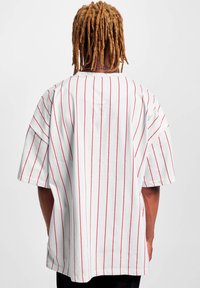 Balta per didelė t-shirt su raudonomis vertikaliomis juostelėmis, trumpomis rankovėmis ir apvaliu kaklu. Struktūruotas medvilnės audinys, laisvas kirpimas.