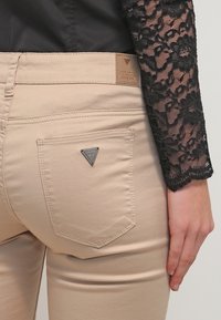 Beiga tajta jeans med en bakficka som har en triangulär svart "Guess"-logotyp. Kombineras med en svart spetslångärmad topp.