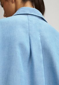 Femme avec une queue de cheval portant une chemise bleu clair à col, vue de dos, mettant en avant la texture du tissu et le détail des coutures.