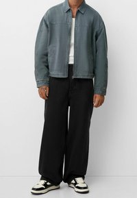 Person som bär en grå zip-up jacka, vit linne, vida svarta byxor och svart-vita sneakers står mot en enfärgad bakgrund.