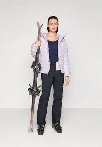 Svetlo vijolična prešita jakna, temno modra majica, črne smučarske hlače, drži smuči z vezmi, obuti v črne smučarske škornje, nevtralno ozadje.