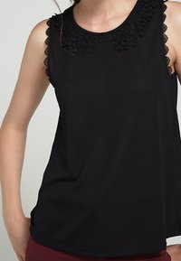 Černý bezrukávový top s krajkovým květinovým detailem kolem výstřihu, hladkým materiálem a uvolněným střihem s zaobleným lemem.