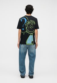 Hombre con cabello rizado que lleva una camiseta negra con una gran cara de dibujos animados verde y azul y un signo de interrogación en la espalda, combinado con pantalones vaqueros azules holgados y zapatos negros.