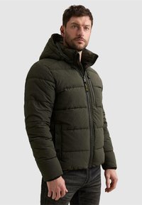 Homme portant une veste matelassée à capuche vert foncé avec fermeture éclair sur le devant et pantalon noir sur un fond clair uni.