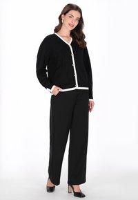 Zwarte cardigan met witte bies en knopen, gecombineerd met zwarte broek met brede pijpen. Soepele stof, V-hals ontwerp en zakken in de cardigan.