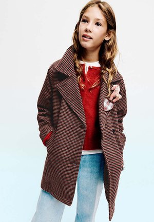 Fille aux longs cheveux ondulés porte un manteau à carreaux marron par-dessus un pull rouge et un jean bleu clair, les mains dans les poches du manteau.