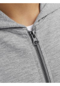 Sweat à capuche gris zippé présente une surface lisse, une capuche texturée, et un détail de fermeture éclair noire robuste le long de l'ouverture frontale.