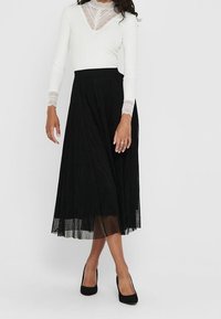 Jupe midi plissée noire avec une texture douce et fluide, associée à un haut à manches longues blanc avec un détail en dentelle au niveau du col et des manches ajustées.