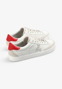 Zapatillas blancas de lona bajas con cordones beiges, acentos rojos en el talón y un emblema de calavera y huesos cruzados bordado en el costado.