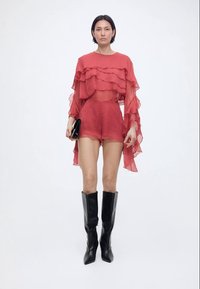 Γυναίκα που φοράει διαφανές κόκκινο μπλουζάκι με στρώσεις και αντίστοιχα σορτς, κρατάει μαύρο τσαντάκι χειρός και φοράει μαύρες μπότες μέχρι το γόνατο, στέκεται μπροστά σε λευκό φόντο.