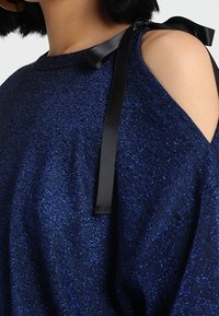 Tummansininen neulepaita, jossa on tekstuuri neuloksessa, kylmä olkapää -malli ja musta satiininauha olkapäillä.